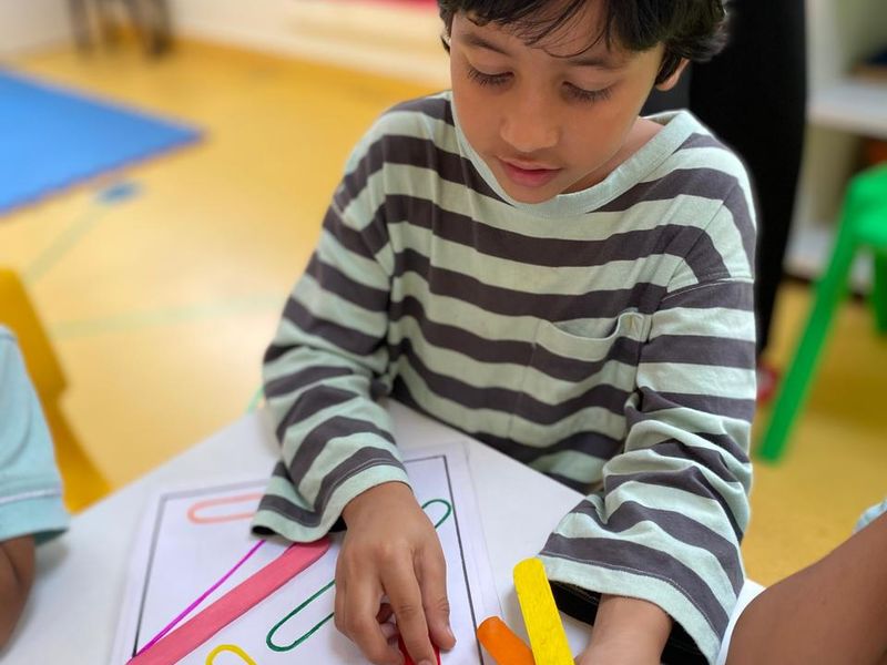An LKG child copying a pattern with coloured craft sticks